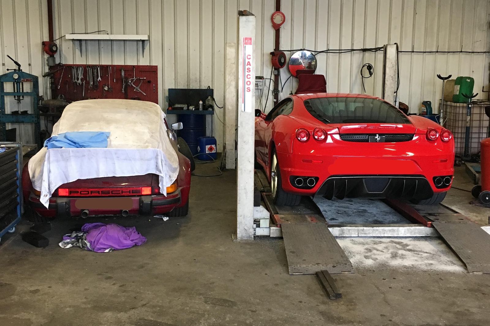 Une Ferrari F430 rouge sur le pont de l’atelier, à côté d’une Porsche 911 couverte d’une housse